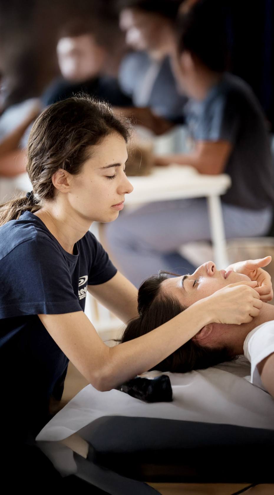 Im Vordergrund ist eine Frau mit dunklem Pferdeschwanz zu sehen, die eine andere Frau auf einer weißen Behandlungsliege behandelt. Die behandelnde Person trägt ein dunkelblaues T-Shirt und hat geschlossene Augen. Sie hält mit beiden Händen den Kopf der liegenden Frau, deren Augen ebenfalls geschlossen sind. Die Patientin liegt auf dem Rücken mit dem Kopf auf einem weißen Polster. Im Hintergrund sind mehrere weitere Personen an ähnlichen Liegen zu erkennen, jedoch unscharf dargestellt. Die Szene findet in einem gut beleuchteten Raum mit hohem Kontrast zwischen Vorder- und Hintergrund statt.